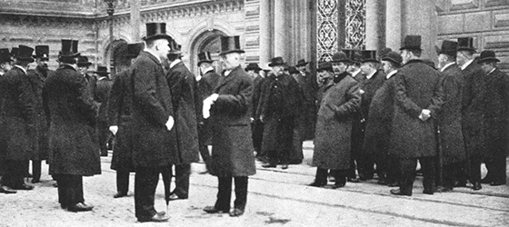 Ett svartvitt fotografi av män som står på gatan och har klädd sig med mörka kläder och cylinderhattar.